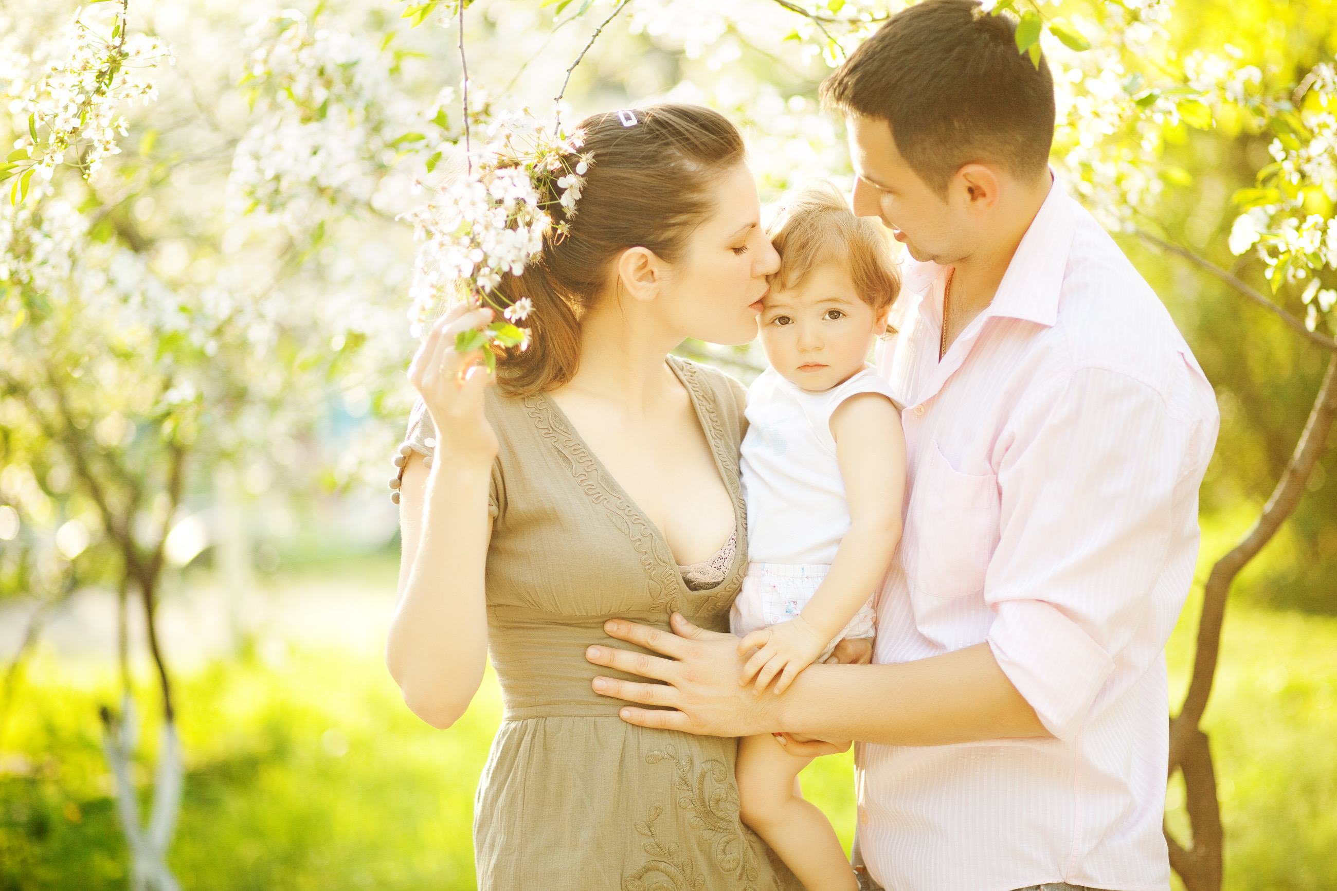 A mother kissing her baby's forehead in a blossoming orchard, the father beside them